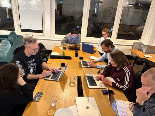 A group of people are gathered around a long wooden table in an indoor office setting, focused on their open laptops.