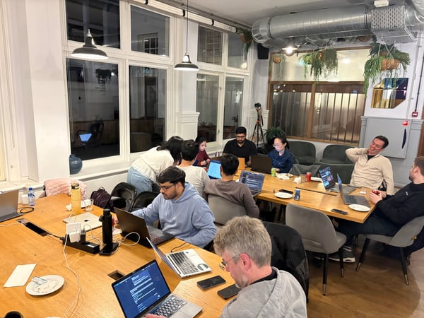 A group of people are gathered around a long wooden table in a modern office or coworking space, focused on their laptops.