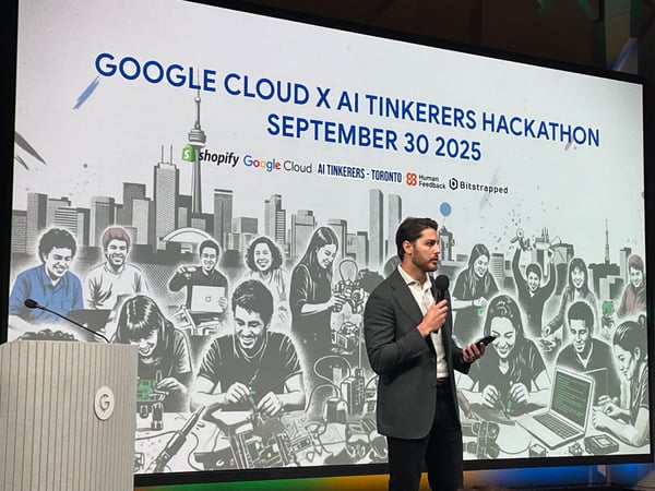 A photograph captures a man speaking into a microphone on stage during a conference, standing in front of a large screen displaying the title for the 'Google Cloud X AI Tinkerers Hackathon' scheduled for September 30, 2025.