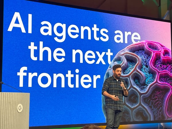 A man is giving a presentation on stage in front of a large screen that displays the text, "AI agents are the next frontier," next to a colorful, abstract, brain-like graphic.