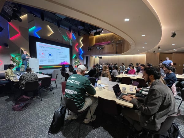 A photograph capturing a technical presentation taking place in a modern auditorium with a large audience seated at tables, many using laptops. A speaker stands on the stage next to a screen displaying a slide about 'AI Field Architecture @ Google'.