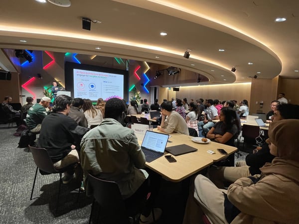 A photograph capturing a large audience attending a presentation in a modern, dimly lit conference room or auditorium, featuring a speaker on a stage in front of a large screen displaying a slide about AI Field Architecture.