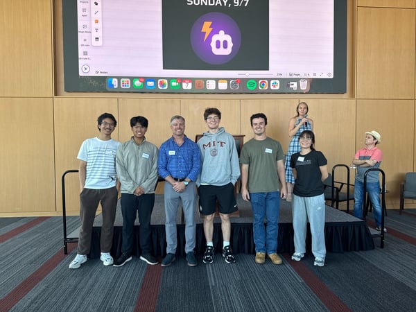 A group of seven individuals, likely participants and organizers of an event, are posing on a small stage in front of a large projection screen displaying the date and a logo.