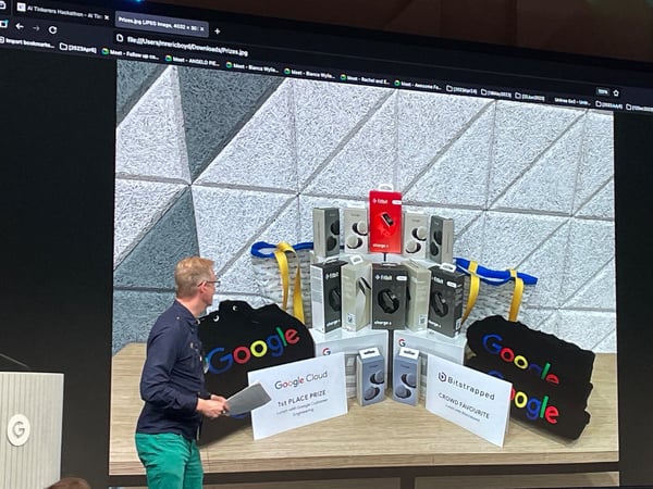 A photograph taken during an event presentation, showing a male speaker standing beside a podium while a large screen displays an image of hackathon prizes, including Fitbit devices and Google merchandise.