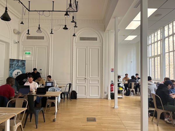 A candid photograph taken inside a large, ornate room with white walls and high ceilings, showing a group of people working on laptops at long tables, suggesting a co-working session or hackathon.