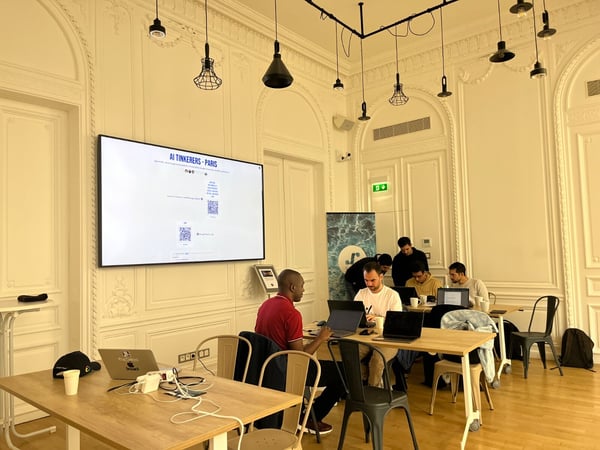 A photograph capturing an indoor workshop or meeting taking place in an elegant room featuring ornate white wall paneling and modern industrial lighting fixtures. Several men are seated at wooden tables, actively working on laptops, while a large screen displays information for an event titled "AI TINKERERS - PARIS".