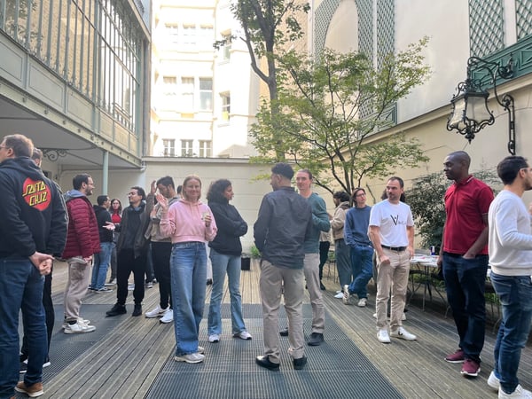 A casual group of people, appearing to be colleagues or attendees of an event, are gathered in an outdoor courtyard setting, standing on a wooden deck surrounded by traditional architecture. They are engaged in conversation and socializing.