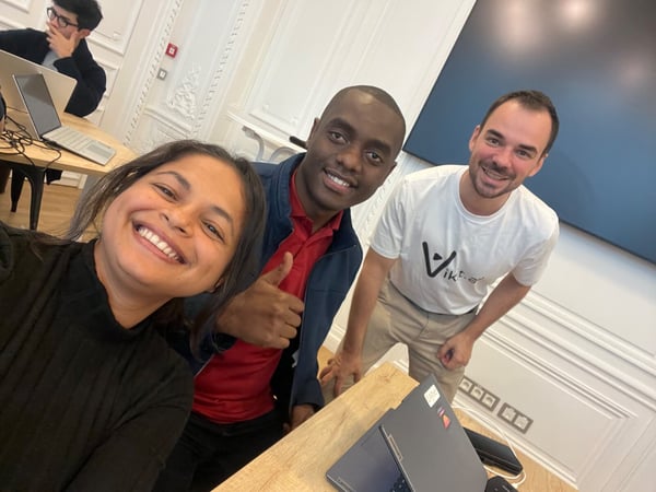 A casual, close-up photograph (likely a selfie) showing three smiling colleagues gathered around a desk in a bright office environment, with one person giving a thumbs-up gesture.