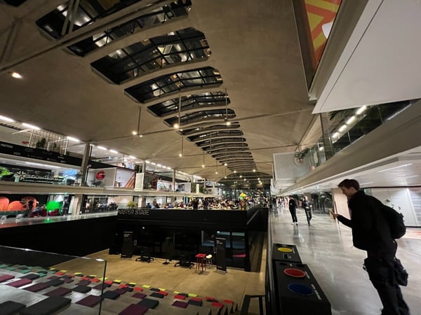A low-angle photograph captures the interior of a large, multi-story venue, likely an event or conference hall, featuring industrial architecture, sponsor displays, and a central area labeled "MASTER STAGE" with tiered cushion seating.