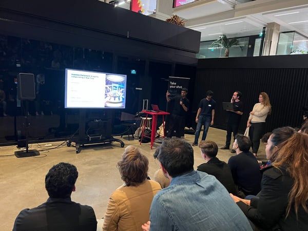 A photograph capturing an indoor presentation event where four individuals stand near a large screen displaying a slide about "Invention Parliament: AI Agents Collaborating in Real Time," while an audience watches from the foreground.