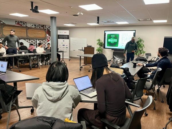 A photograph capturing an indoor workshop or presentation session where a speaker addresses a group of attendees seated at tables with open laptops, while a large screen displays a graphic featuring the text "WORLDWIDE INNOVATION."