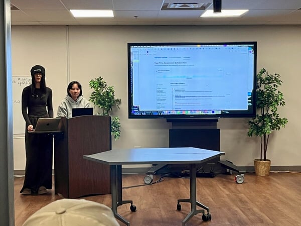 A photograph captures two women giving a presentation or demonstration in a modern conference room, standing near a podium in front of a large screen displaying a user interface titled "Real-Time Negotiation Collaboration."