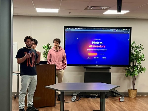 A photograph capturing three young men in an indoor meeting or presentation setting, where one man stands near a podium looking at his phone, and a large screen behind them displays a presentation slide titled "Pitch to AI Investors."