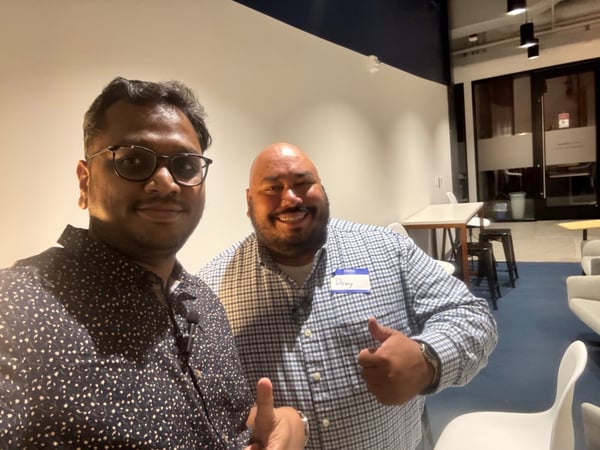 A selfie featuring two men smiling and giving thumbs up gestures indoors, likely taken at a networking event or office social gathering.