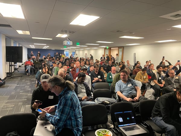A large group of people are seated in rows of chairs in a modern office or conference room, many of whom are looking at their phones or laptops.