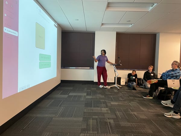 A man in a purple t-shirt and red plaid pants is giving a presentation to a small audience in an office setting using a projector screen.
