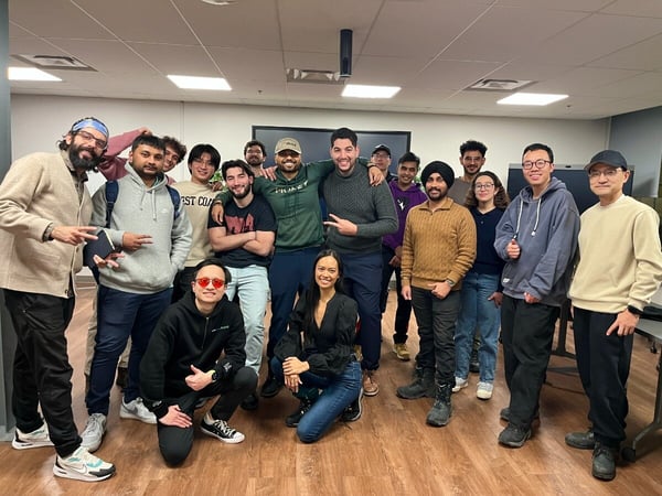 A large diverse group of approximately seventeen people poses closely together for a casual group photo indoors, likely in an office or meeting room setting with wood flooring and fluorescent lighting.