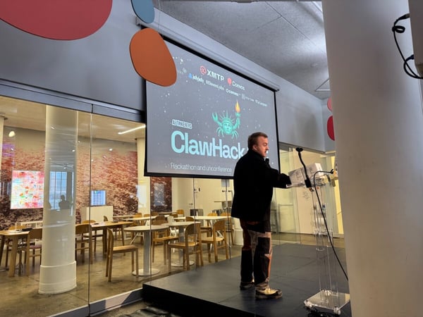 A man stands on a stage in an indoor event space, positioned in front of a projection screen displaying 'ClawHack: Hackathon and unconference.'