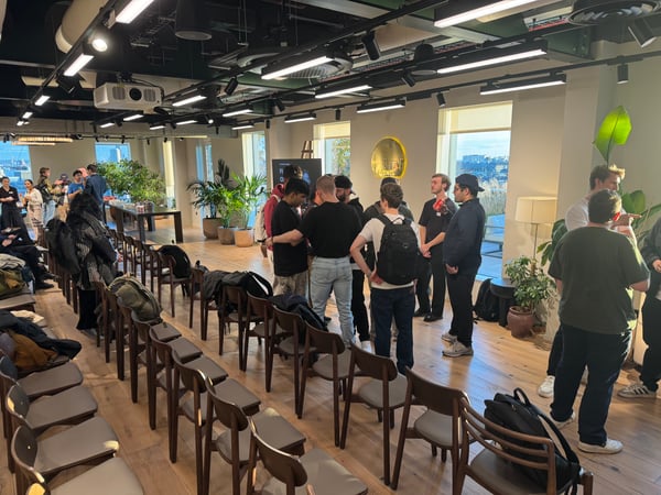 A candid photograph taken inside a modern, well-lit office or event space, showing a group of people mingling and networking around the center of the room. Rows of mostly empty chairs are arranged in the foreground, suggesting the space is set up for a presentation or meeting.