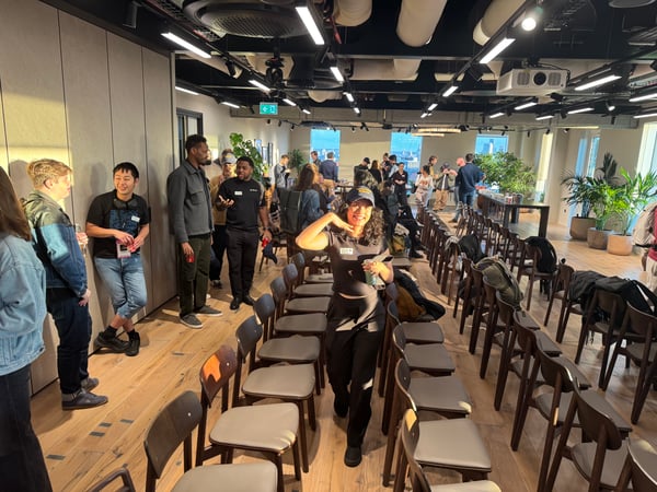 A candid photograph capturing a group of people socializing and networking in a modern, brightly lit event space set up with rows of empty chairs for an audience. A woman in the foreground smiles while looking directly at the camera.