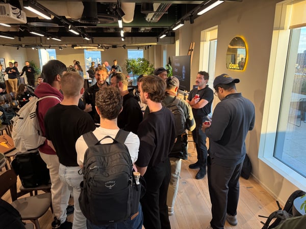 A candid photograph captures a large group of people, mostly men, gathered indoors in a modern office space, likely networking after an event. A screen displaying 'Fireside Chat' and an illuminated sign reading 'dawn.' are visible on the far wall.