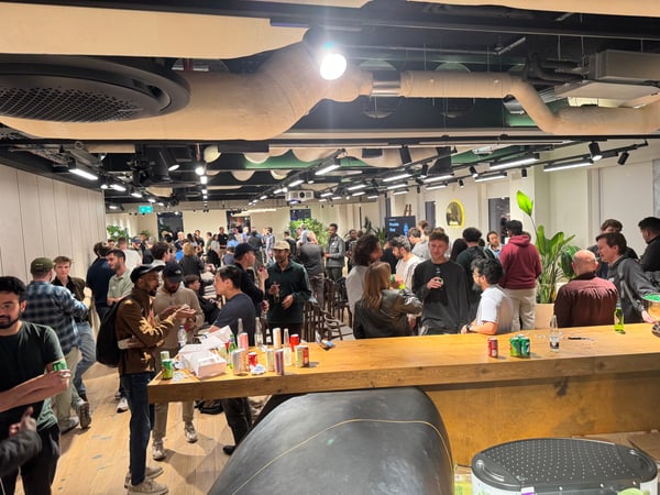A candid photograph capturing a large group of people mingling and socializing around a wooden bar or counter at a crowded indoor event, likely a networking gathering in a modern office space.