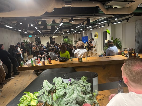 A candid photograph captures a large audience gathered indoors, listening to two speakers seated on high stools on a small stage area under low, atmospheric lighting. The foreground features a wooden bar counter topped with potted plants and various beverage containers.