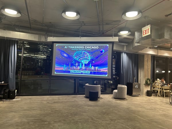 A photograph capturing the setup of an indoor event space at night, featuring a large screen displaying a colorful slide for the 'AI Tinkerers Chicago' meetup, set against a backdrop of a city skyline visible through large windows.