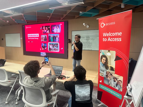 A photograph capturing an indoor corporate presentation where a male speaker is addressing an audience in front of a large screen displaying a slide titled "Introduction to The Access Group" and a prominent red 'Welcome to Access' roll-up banner.