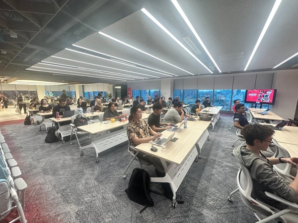 A wide shot of a crowded indoor event, likely a workshop or training session, where dozens of participants are seated at long tables with laptops open in a brightly lit room with large windows overlooking a city skyline.