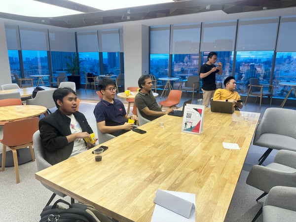 A photograph showing five men gathered around a large wooden table in a modern, high-rise office lounge area, seemingly attending a casual presentation or meeting while overlooking a city skyline at dusk.