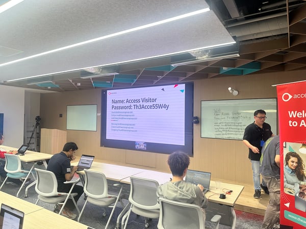 A photograph taken inside a modern training room or office showing several attendees working on laptops while an instructor stands near a whiteboard and a large screen displaying Wi-Fi credentials and contact emails for 'ACCESS'.