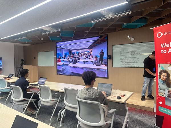 A photograph of an indoor training session or workshop taking place in a modern room, showing participants working on laptops while facing a large video screen that displays a remote audience or class.
