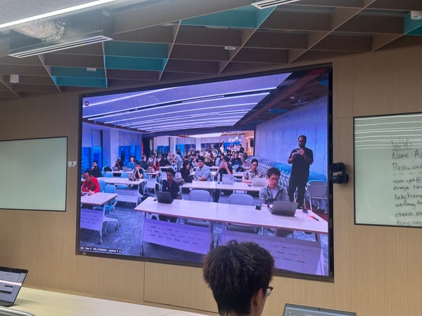A photograph taken inside a modern classroom or meeting room, showing a large video wall displaying a live feed or recording of a lecture hall filled with students and a presenter.