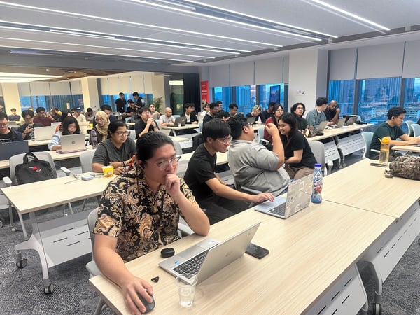 A wide-angle photograph capturing a large group of people attending a workshop or training session in a modern, brightly lit office space. The attendees are seated at long tables, mostly focused on their open laptops, with a city skyline visible through the large windows in the background.