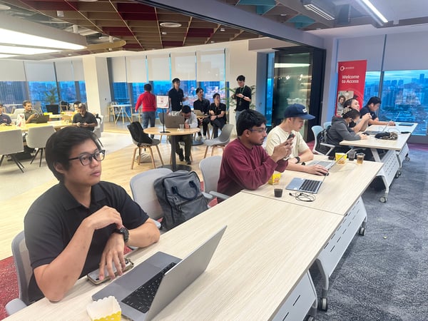 A photograph captures a group of people attending a focused session or workshop inside a modern, well-lit office space, featuring large windows overlooking a city skyline at dusk. Attendees are seated at long tables, many using laptops and mobile phones, facing a common direction off-camera.
