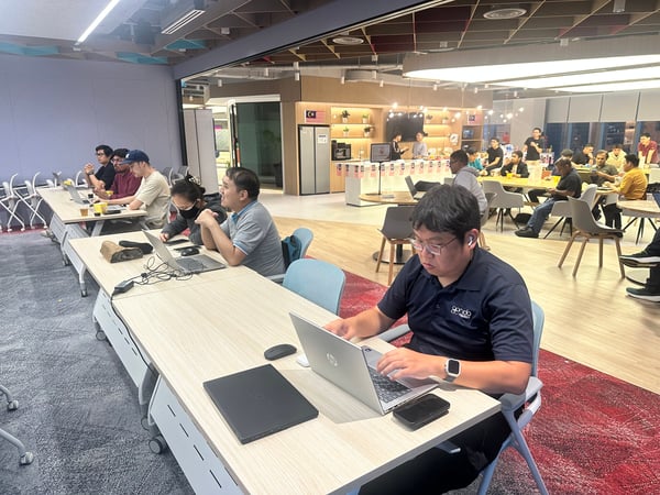 A photograph capturing a group of people working intently on laptops at long tables in a modern, brightly lit office or co-working space during what appears to be a focused work session or training event.