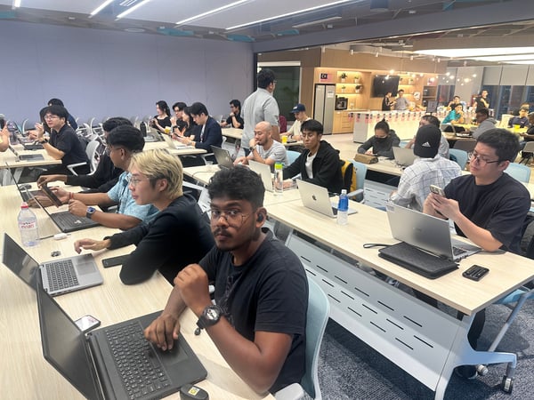 A candid photo taken indoors showing a large group of people, primarily young adults, seated at long tables and working intently on their laptops and mobile phones during a workshop or coding session.
