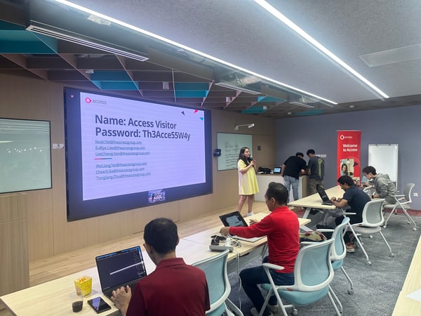 This image captures an indoor corporate training or presentation session where a woman is speaking into a microphone in front of a large screen displaying Wi-Fi login details and company email addresses. Several attendees are seated at tables, actively using their laptops.