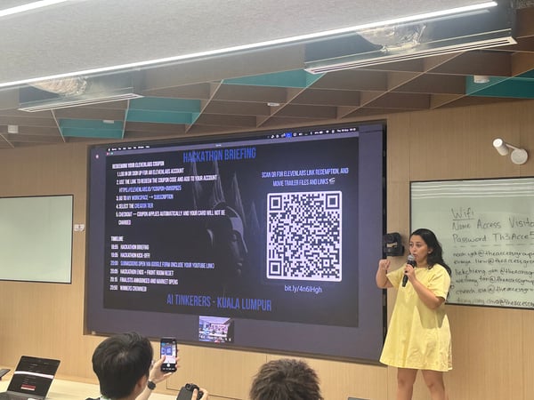 A photograph capturing an indoor event where a woman in a yellow dress is giving a presentation, holding a microphone, in front of a large screen displaying a 'Hackathon Briefing' with instructions, a timeline, and a QR code. Several audience members are visible in the foreground, some taking pictures with their phones.