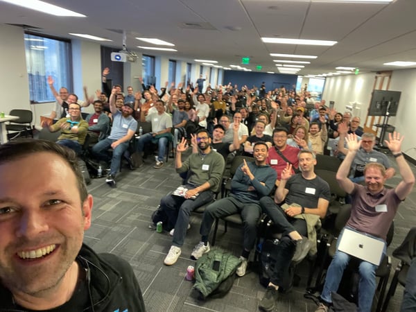 A wide-angle group selfie taken by a smiling man in the foreground, showing a large crowd of people seated in an office or presentation room waving at the camera.