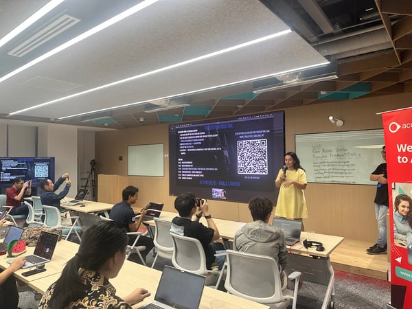 A photograph captures a technical presentation or briefing taking place in a modern office classroom setting. A woman in a yellow dress is speaking into a microphone on a small stage while an audience, seated at tables with open laptops, watches the large screen displaying a QR code and instructions.