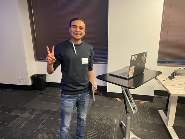 A young man stands smiling in an office setting, making a peace sign with his hand next to a laptop on a standing desk.