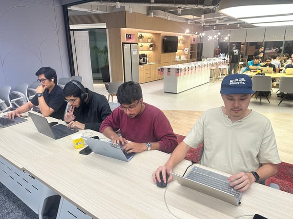 Four young men are seated at a long wooden table, intensely focused on working on their laptops in a modern, well-lit office or co-working space decorated with Malaysian flags.