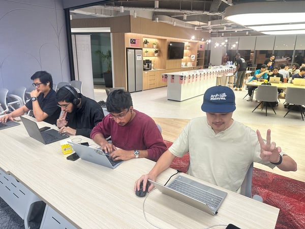 A candid photograph showing a group of young men working intensely on laptops at a long desk in a modern, well-lit office or co-working space, possibly during a hackathon. The man in the foreground is wearing a cap that reads "Ayuh Malaysia." and is making a peace sign.