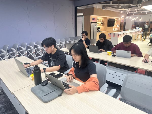 A candid photograph captures several young adults sitting at light wood desks, deeply focused on working on their individual laptops in a modern indoor setting. The room appears to be a training or co-working space, with Malaysian flags visible in the communal area in the background.