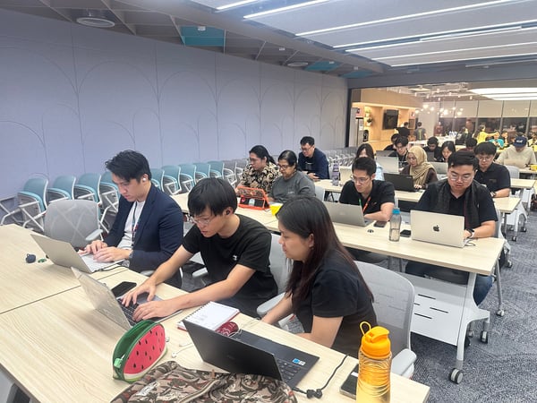 A photograph showing a diverse group of people seated at long tables in a modern office or training room, intensely focused on working on their open laptops.