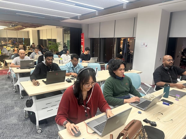A candid photograph showing a diverse group of approximately eighteen people working intently on open laptops at long desks in a brightly lit, modern office environment.