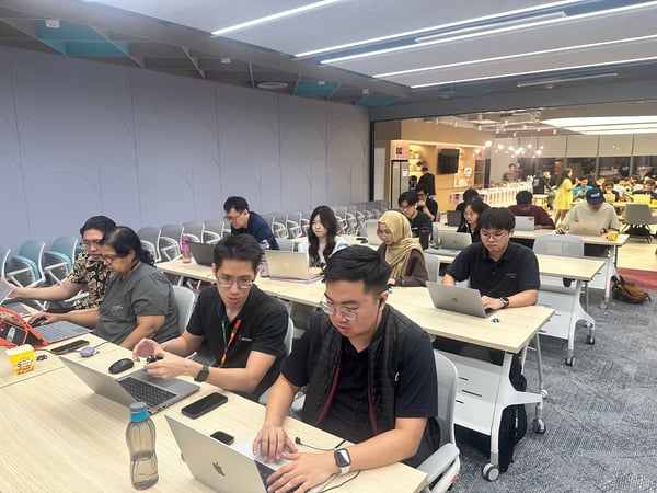 A photograph showing a diverse group of people sitting at long tables in a modern, brightly lit indoor workspace, intently working on their individual laptops during what appears to be a coding session or hackathon.
