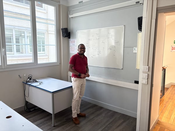A man stands smiling in a room next to a whiteboard filled with technical diagrams and notes.